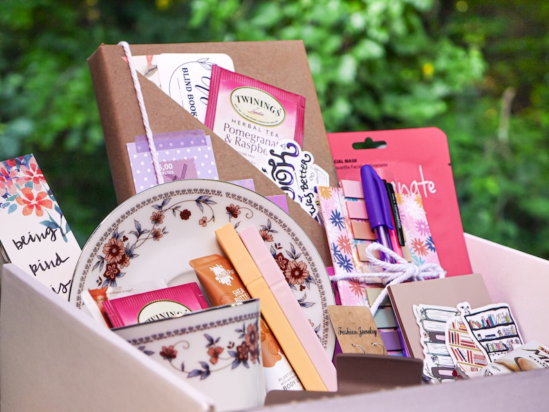 May include: A pink and white gift box filled with various items, including a brown notebook, a tea bag, a pink and purple sticker, a floral patterned pouch, a purple pen, a pink and white patterned notepad, a pink and white patterned bookmark, a teacup and saucer, and a pink and white patterned sticker. The box is surrounded by green foliage.