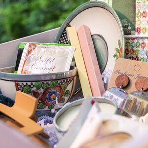 May include: A gift box filled with various items, including a teacup and saucer, a candle, a hair clip, a package of salted caramel creamer, and a pair of wooden earrings with a book-themed design. The box is lined with white tissue paper and has a light pink background.