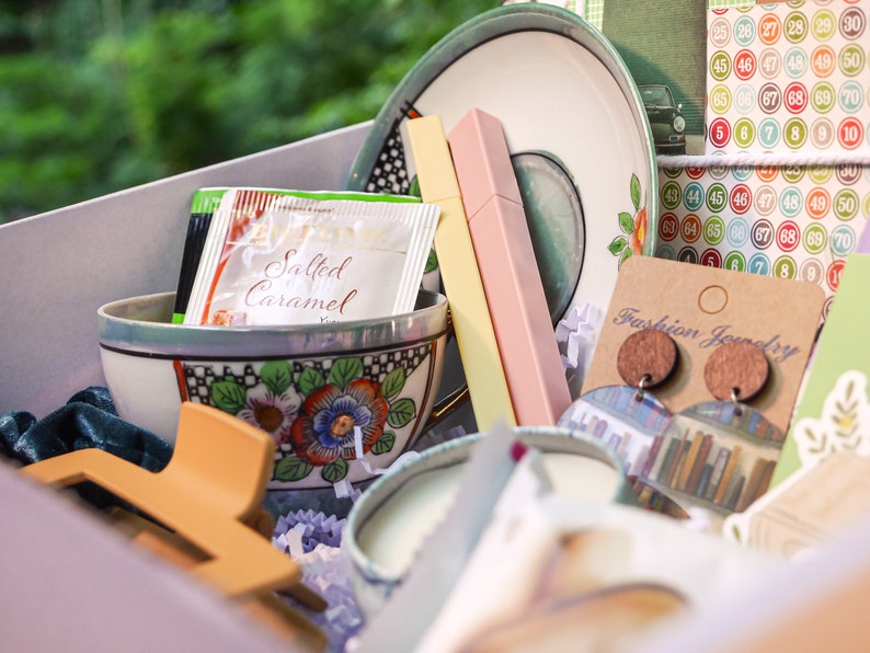 May include: A gift box filled with various items, including a teacup and saucer, a candle, a hair clip, a package of salted caramel creamer, and a pair of wooden earrings with a book-themed design. The box is lined with white tissue paper and has a light pink background.