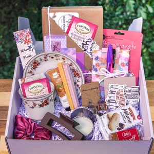 May include: A purple and white gift box filled with various items, including tea, bath products, jewelry, and stationery. The box is filled with purple and white tissue paper. The items are arranged in a visually appealing way.