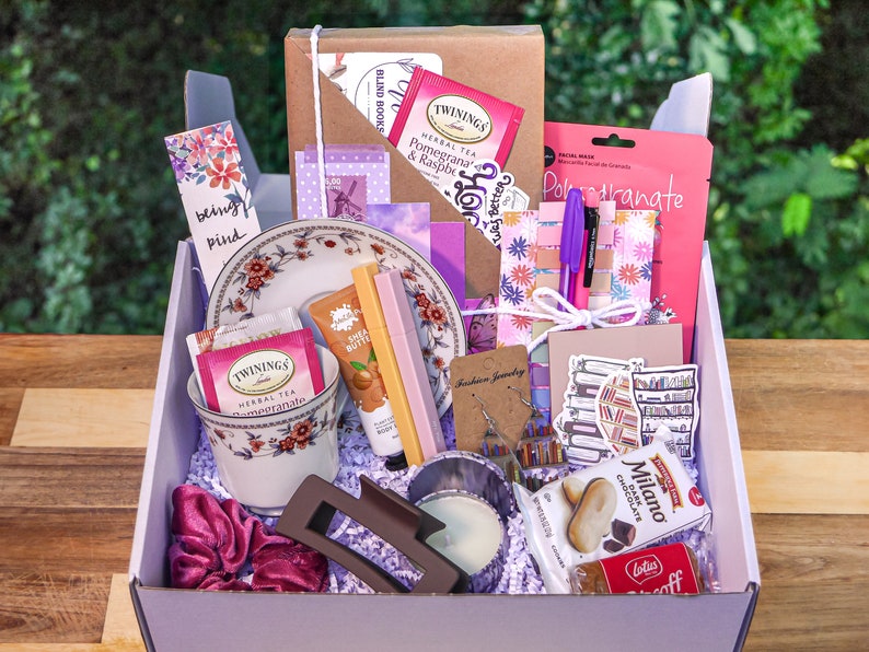 May include: A purple and white gift box filled with various items, including tea, bath products, jewelry, and stationery. The box is filled with purple and white tissue paper. The items are arranged in a visually appealing way.