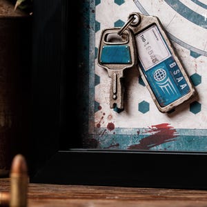 May include: A framed artwork featuring three keys. One key has a blue accent, and another has a tag that reads "BSAA" and "THD-19528." The background has a map design with red and blue accents. The artwork is in a black frame.