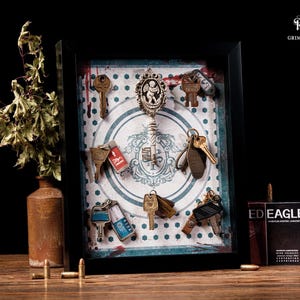 May include: A framed display featuring antique keys and a decorative key pendant. The keys are arranged against a patterned background with a vintage aesthetic. A small box of ammunition and a vase with dried foliage are also present.