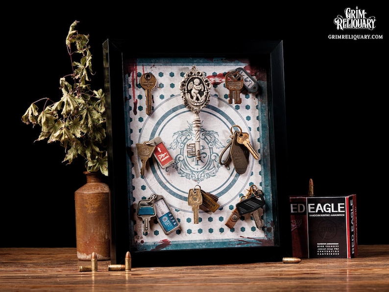 May include: A framed display featuring antique keys and a decorative key pendant. The keys are arranged against a patterned background with a vintage aesthetic. A small box of ammunition and a vase with dried foliage are also present.