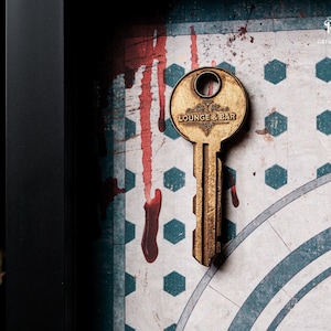 May include: A vintage-style key with "LOUNGE & BAR" text, set against a backdrop of a patterned surface with red streaks. The key is gold-toned, and the background has a geometric design with blue and white shapes.