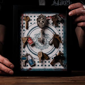 May include: A framed display featuring various antique keys and keychains. The keys are arranged on a patterned background with a central decorative element. The frame is black, and the overall aesthetic is vintage and gothic.