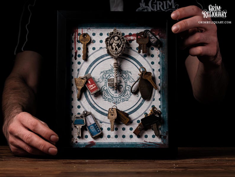 May include: A framed display featuring various antique keys and keychains. The keys are arranged on a patterned background with a central decorative element. The frame is black, and the overall aesthetic is vintage and gothic.