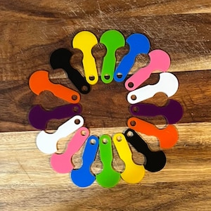 May include: A set of 15 colourful plastic bottle openers arranged in a circle on a wooden surface. Each opener has a unique colour and a different shape.
