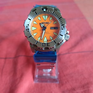 May include: A Seiko automatic dive watch with a stainless steel band and case. The watch face is bright orange with luminous markers and hands. The bezel is silver with black markings, and the day and date are displayed at the 3 o'clock position.