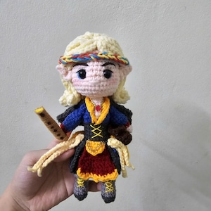 May include: Crocheted doll with blonde hair, wearing a blue and red dress with a yellow belt and a brown bag. The doll is holding a wooden flute.