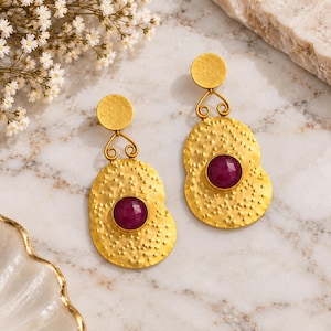 May include: A pair of gold-toned drop earrings featuring a textured, hammered finish on the main oval-shaped component. Each earring is accented with a single, round, faceted deep magenta gemstone set in the center and a small circular stud top.