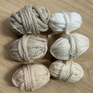 Könnte beinhalten: Sechs Knäuel Garn in verschiedenen Beige- und Weißtönen. Das Garn ist in zwei Reihen mit jeweils drei Knäueln angeordnet.