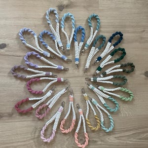 Könnte beinhalten: Eine Sammlung von 18 bunten gehäkelten Schlüsselanhängern mit weißen Akzenten. Die Schlüsselanhänger sind in einem Kreis auf einer Holzoberfläche angeordnet.