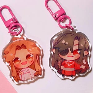 May include: Two acrylic keychains featuring chibi-style anime characters with pink plastic lobster clasps. The left character has light brown hair and wears white clothing. The right character has dark hair, an eyepatch, and a red outfit.
