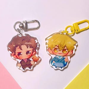 May include: Two acrylic keychains featuring chibi-style anime characters. The left keychain has a silver clasp and a character with brown hair in a suit holding a gavel. The right keychain has a yellow clasp and a character with blonde hair in blue.