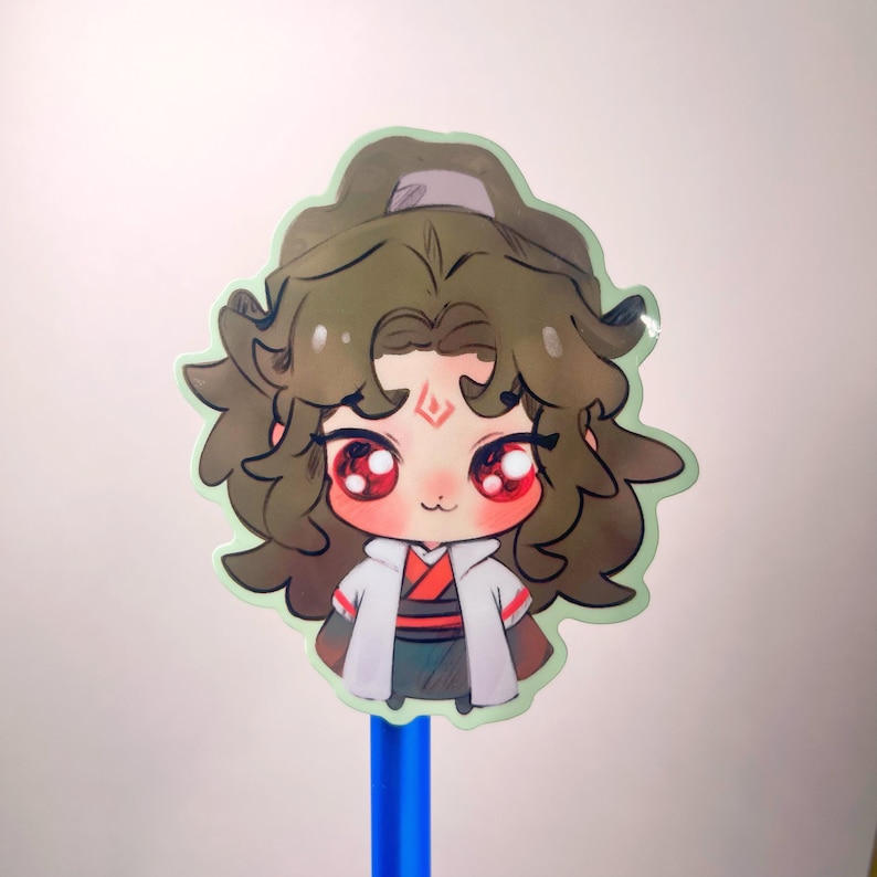 May include: A sticker of a cartoon character with brown hair, red eyes, and a white and red outfit. The sticker is attached to a blue pen.