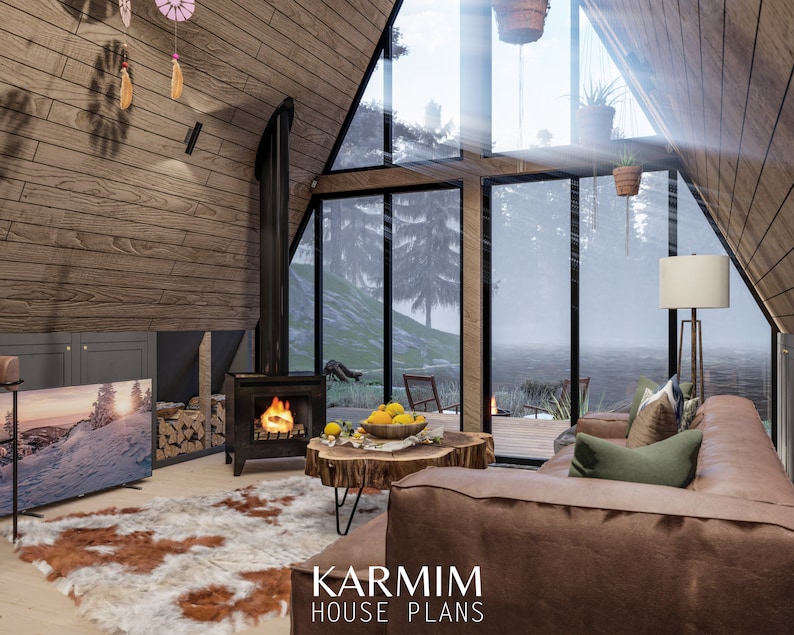 May include: A cozy living room with a large window overlooking a snowy forest. The room features a wood-burning stove, a leather sofa, a coffee table, and a television. The text "KARMIM HOUSE PLANS" is visible in the bottom right corner of the image.