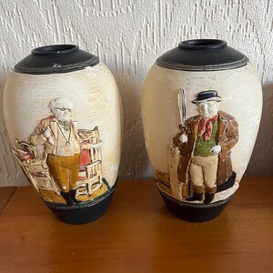 May include: Two decorative ceramic vases with black tops and cream-colored bodies. Each vase features a raised relief of a person in vintage clothing. One vase depicts a man with glasses, the other a man with a hat.