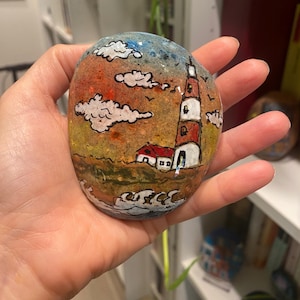 May include: Hand-painted rock art depicting a lighthouse with a red roof and white stripes, a small house, and waves crashing against the shore. The background features a sunset with orange, yellow, and blue clouds.