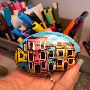 May include: Hand-painted rock art depicting a row of colorful houses with windows and doors. The houses are painted in shades of yellow, pink, blue, and green. The rock is painted with a blue sky and white clouds.