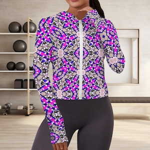 Puede incluir: Chaqueta deportiva con cremallera con un vibrante estampado geométrico en tonos rosa, morado, negro y blanco. La chaqueta tiene mangas largas y una cremallera blanca. La persona lleva leggings grises oscuros.