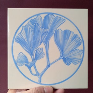 Könnte beinhalten: Eine weiße Keramikfliese mit einem blauen Ginkgoblatt-Design. Die Blätter sind stilisiert und in einem kreisförmigen Muster angeordnet.