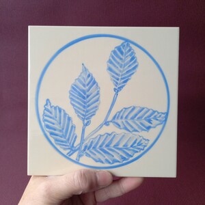Peut inclure: Un carreau de céramique blanc avec un motif circulaire bleu représentant des feuilles bleues stylisées. Les feuilles sont disposées en cercle et ont une apparence texturée.
