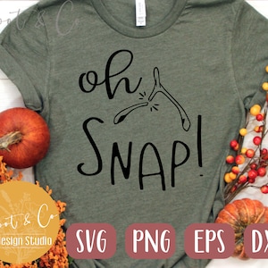 May include: A green t-shirt with the text "oh snap!" in black lettering. The text is accompanied by a graphic of a wishbone breaking. The t-shirt is surrounded by fall-themed decorations, including pumpkins and branches with red berries.