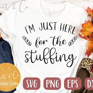 May include: White t-shirt with black text that reads "I'm just here for the stuffing". The text is in a cursive font and is surrounded by two wheat stalks. The shirt is on a wooden background with fall leaves and a pair of leopard print shoes.