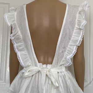 May include: White cotton dress with a deep V-back and ruffled straps. The dress has a tie at the waist and a tiered skirt.