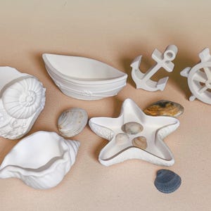 Op de afbeelding: Een verzameling witte nautische decoratieartikelen, waaronder een zeeschelp, een bootvormige schaal, een anker, een scheepsrad, een stervormige schaal en verschillende zeeschelpen. De items zijn gerangschikt op een lichtbeige oppervlak.