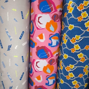 Könnte beinhalten: Drei Stoffrollen mit verschiedenen Mustern. Eine ist grau mit Skateboards, Blitzsymbolen und dem Wort "TOUR". Eine andere ist rosa mit abstrakten Formen in Blau, Orange und Weiß. Die dritte ist blau mit gelben Blumen.