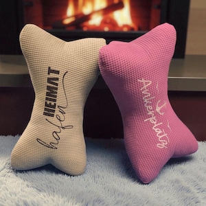 Könnte beinhalten: Zwei dekorative, knochenförmige Kissen. Eines ist beige mit den Wörtern "HEIMAT" und "hafen" bedruckt. Das andere ist rosa mit dem Wort "Ankerplatz" bedruckt. Die Kissen liegen auf einer blauen, flauschigen Oberfläche, mit einem Kamin im Hintergrund.