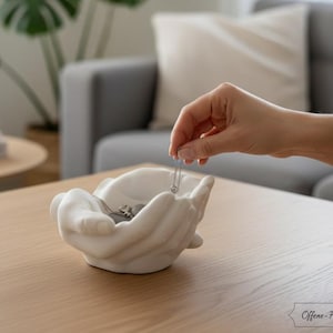 Könnte beinhalten: Eine weiße Keramiktasse mit schwarzem Henkel und dem Text "But First, Coffee" in fetter, schwarzer Schrift. Die Tasse steht vor einem verschwommenen Hintergrund, der eine gemütliche Umgebung suggeriert.