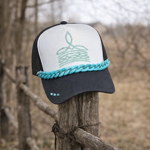 May include: A black trucker hat with a white front panel featuring a teal design. The brim is black, and a teal chain detail accents the hat. Three teal gems are on the brim. The hat is resting on a wooden post.