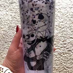 May include: A clear tumbler with a black and white illustration of Jack Skellington and Zero from The Nightmare Before Christmas. The tumbler is filled with glitter and white liquid. The tumbler is held by a person.