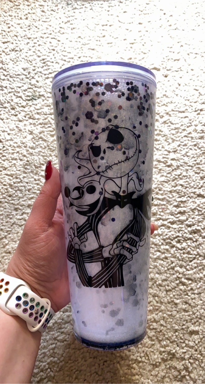 May include: A clear tumbler with a black and white illustration of Jack Skellington and Zero from The Nightmare Before Christmas. The tumbler is filled with glitter and white liquid. The tumbler is held by a person.