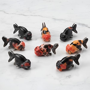 May include: A collection of eight goldfish figurines. The fish are black, orange, and white, with prominent, rounded heads and flowing fins. They are arranged on a white marble surface.