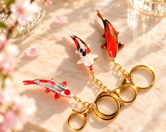 May include: Three koi fish charms attached to gold-toned metal keychains. Each charm features detailed, glossy, hand-painted patterns in red, white, and black. The fish are connected to circular rings and lobster clasps by short, delicate gold-coloured chains.