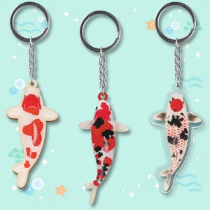 May include: Three koi carp keyrings with silver chain and ring attachments. The fish designs feature red, black, and white patterns. The background is a light blue with decorative bubbles and stars.