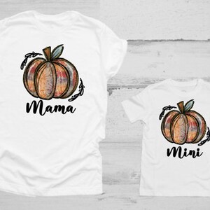Pode incluir: Duas camisetas brancas com um design temático de outono. A camiseta maior tem uma abóbora com a palavra "Mama" embaixo. A camiseta menor tem uma abóbora com a palavra "Mini" embaixo.