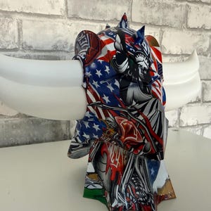 Peut inclure: Une sculpture décorative représentant une tête de taureau stylisée avec de grandes cornes blanches incurvées. La tête est ornée d'un motif vibrant incorporant le drapeau américain, la Statue de la Liberté et d'autres éléments patriotiques. La sculpture est posée devant un mur de briques blanches et grises.