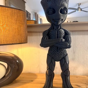 Peut inclure: Figurine noire représentant un personnage humanoïde avec une apparence d'arbre, les bras croisés et une expression souriante. La figurine présente des textures détaillées et se tient sur une surface en bois.
