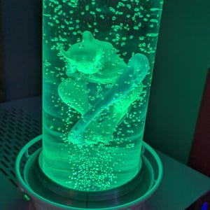 May include: A cylindrical liquid-filled lamp featuring a green-lit interior with a central figurine suspended in the fluid. The lamp has a white base and top cap, with numerous small air bubbles rising through the vibrant green illuminated liquid.