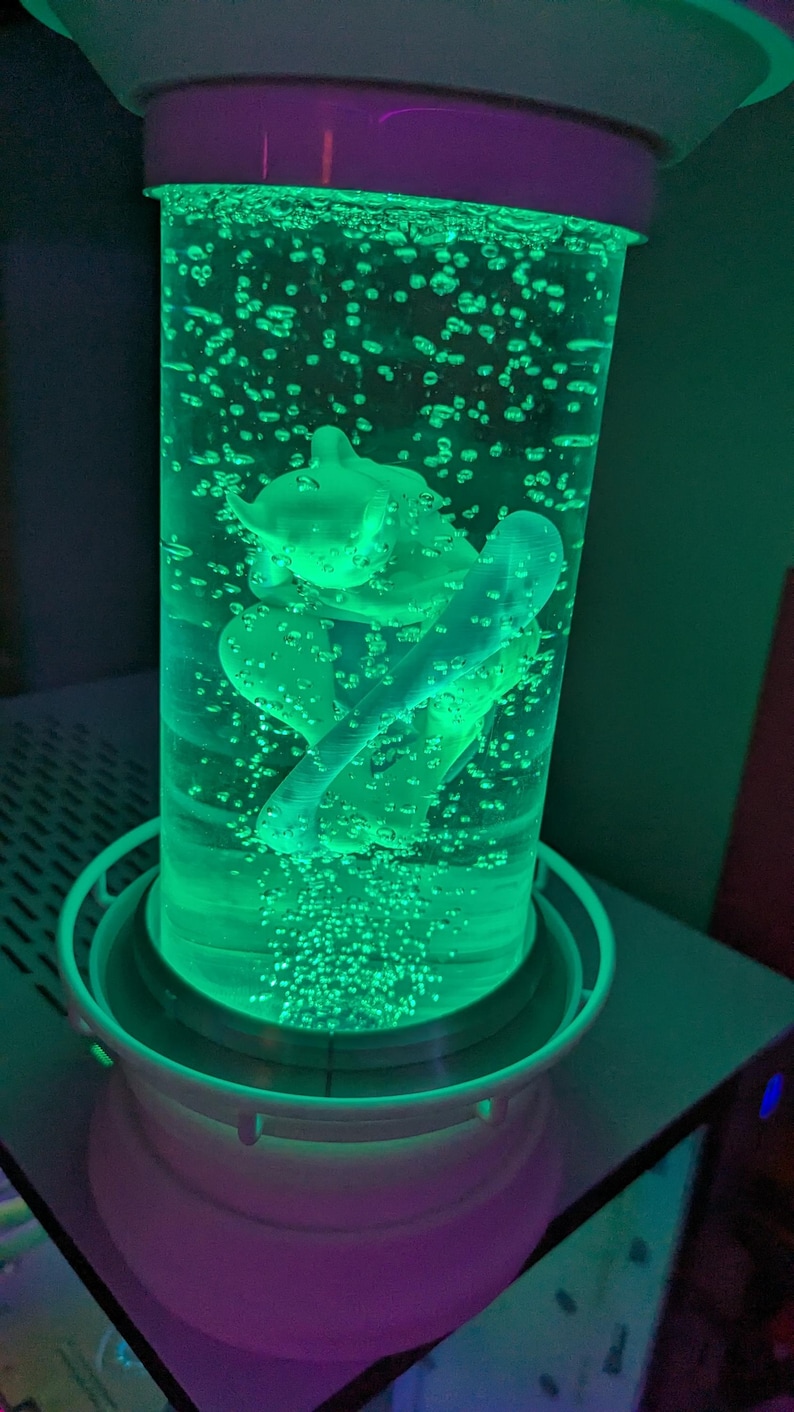 May include: A cylindrical liquid-filled lamp featuring a green-lit interior with a central figurine suspended in the fluid. The lamp has a white base and top cap, with numerous small air bubbles rising through the vibrant green illuminated liquid.