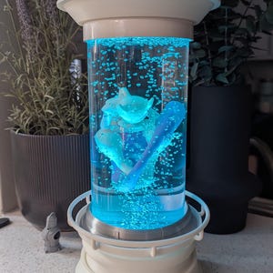 May include: A cylindrical liquid-filled lamp with a white base and top, featuring a bright blue illuminated interior with rising bubbles and a central white figurine. The structure has a mechanical aesthetic with a clear central chamber.
