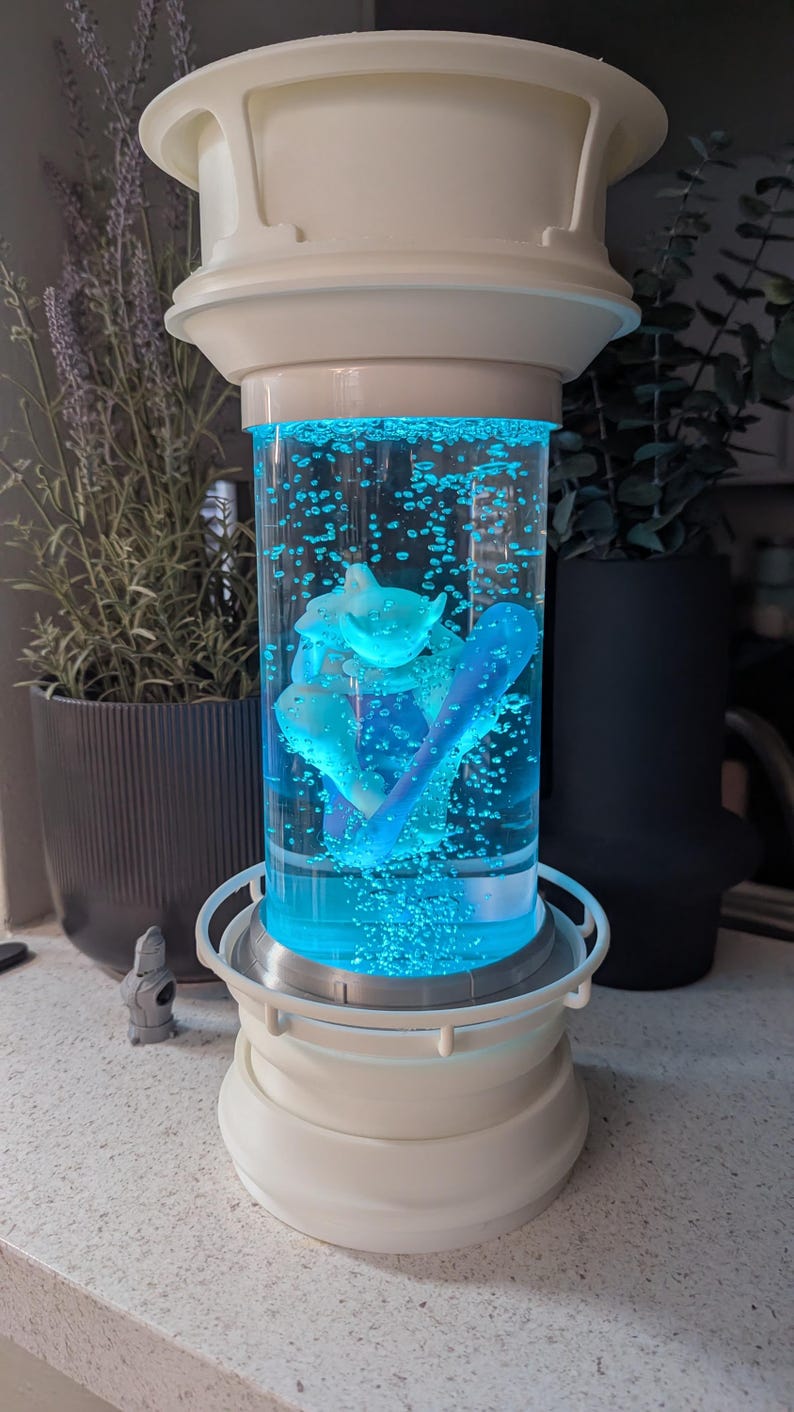 May include: A cylindrical liquid-filled lamp with a white base and top, featuring a bright blue illuminated interior with rising bubbles and a central white figurine. The structure has a mechanical aesthetic with a clear central chamber.