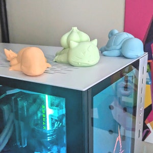 May include: Three 3D printed figurines of cartoon characters, in light blue, light green, and peach, are sitting on top of a black computer case with a glass side panel. The computer case has a blue and green light inside.