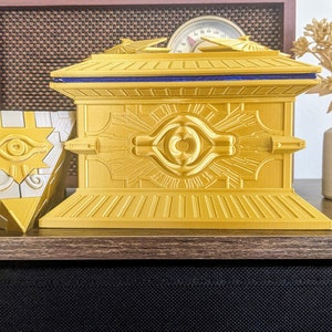 May include: A gold-colored, rectangular box with intricate detailing and a blue accent. Beside it is a gold and silver pyramid-shaped object. The objects are likely decorative or collectible items.