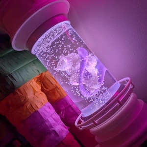 May include: A cylindrical liquid-filled sensory lamp featuring a white base and cap, containing a purple floating shape and numerous small air bubbles. The lamp is illuminated by a vibrant pink neon light, casting a glow on the clear liquid.